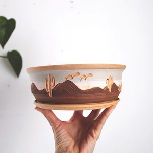 May include: A handmade ceramic planter with a desert landscape design. The brown and beige planter features a painted scene of mountains, cacti, and birds. The planter is held by a hand and includes a matching saucer.