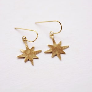 May include: A pair of gold star-shaped earrings with a hammered texture. The earrings are suspended from a simple wire loop.