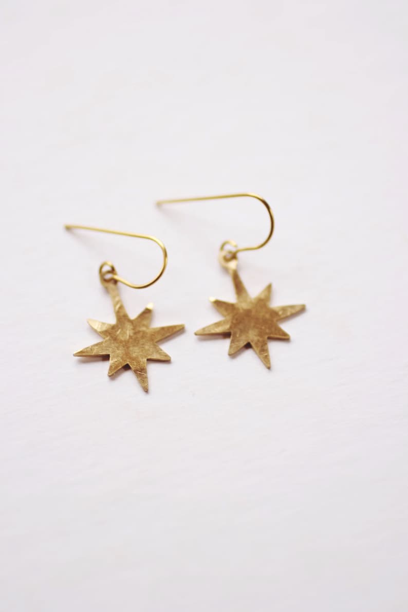May include: A pair of gold star-shaped earrings with a hammered texture. The earrings are suspended from a simple wire loop.