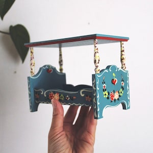 May include: A miniature, hand-painted blue wooden bed with floral and heart details. The bed has a canopy with red trim and decorative posts. The bed is held by a hand against a white background.