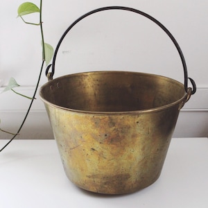 Puede incluir: Un cubo de latón vintage con un asa de metal negro. El cubo tiene una forma ligeramente cónica y muestra signos de edad y uso. El fondo es una superficie blanca lisa.