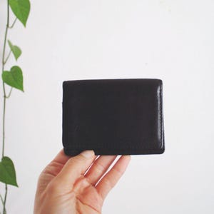 May include: A rectangular dark brown leather wallet with a smooth, matte finish and visible stitched edges. The wallet features a simple, folded design with a clean, minimalist exterior surface and a consistent, dark monochromatic color tone.