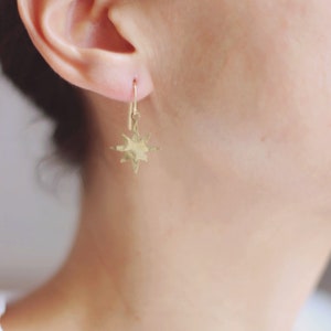 May include: Gold star-shaped earrings with a hammered texture. The earrings are hanging from a simple gold hoop.