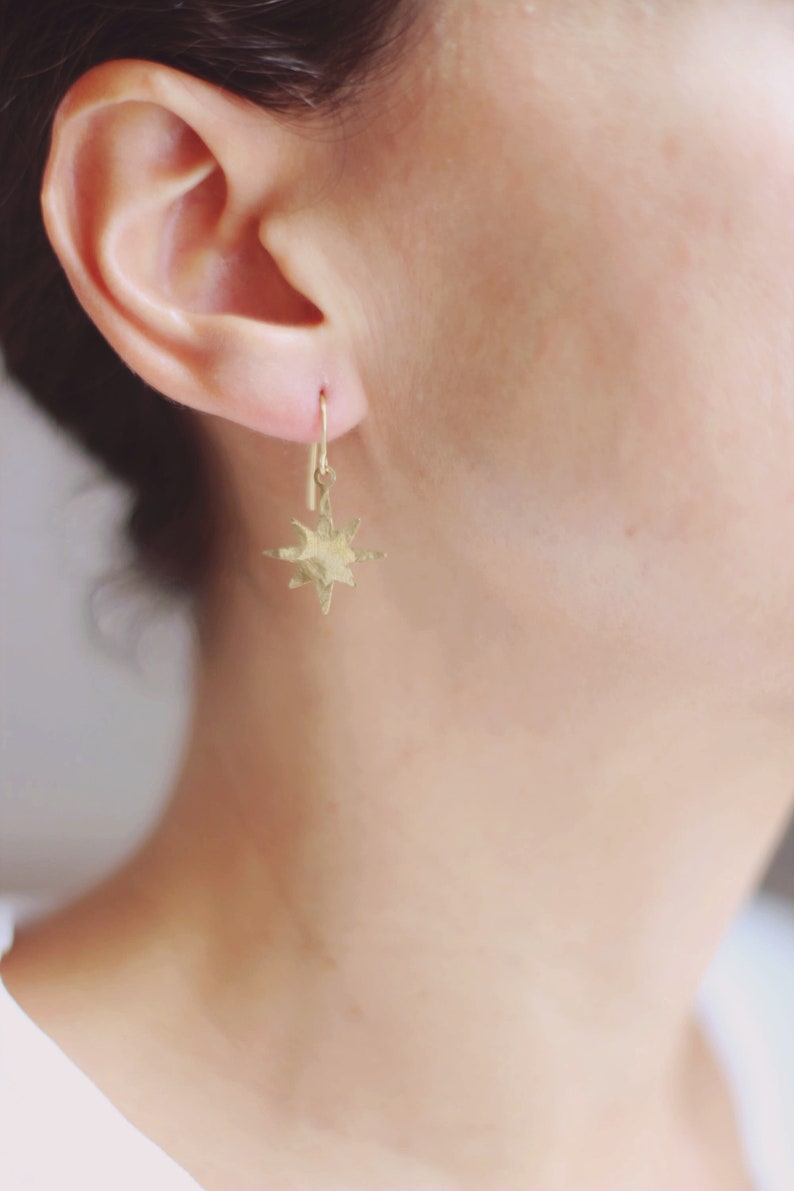 May include: Gold star-shaped earrings with a hammered texture. The earrings are hanging from a simple gold hoop.