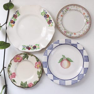 Może przedstawiać: Cztery dekoracyjne ceramiczne talerze z kwiatowymi i owocowymi wzorami. Jeden talerz ma ilustrację brzoskwini, inny niebiesko-białą kratkę, a dwa pozostałe mają wzory kwiatowe w różnych kolorach.