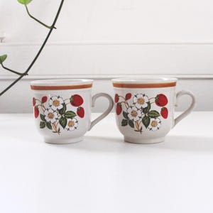 May include: Two ceramic mugs with a cream-colored base, featuring a strawberry and floral design. The mugs have a brown rim and handle, with a repeating pattern of red strawberries and white flowers with green leaves. The mugs are placed on a white surface.