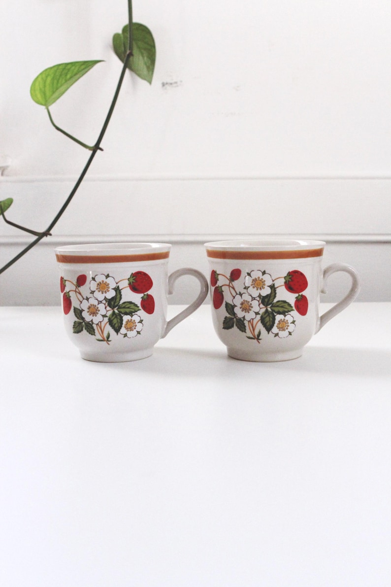 May include: Two ceramic mugs with a cream-colored base, featuring a strawberry and floral design. The mugs have a brown rim and handle, with a repeating pattern of red strawberries and white flowers with green leaves. The mugs are placed on a white surface.