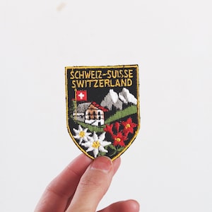 Może przedstawiać: Naszywka w kształcie tarczy z czarnym tłem i ż&oacute;łtym napisem SCHWEIZ-SUISSE SWITZERLAND. Wz&oacute;r przedstawia małą szwajcarską flagę, g&oacute;rski domek, ośnieżone szczyty, białe kwiaty szarotki oraz czerwone kwiaty.