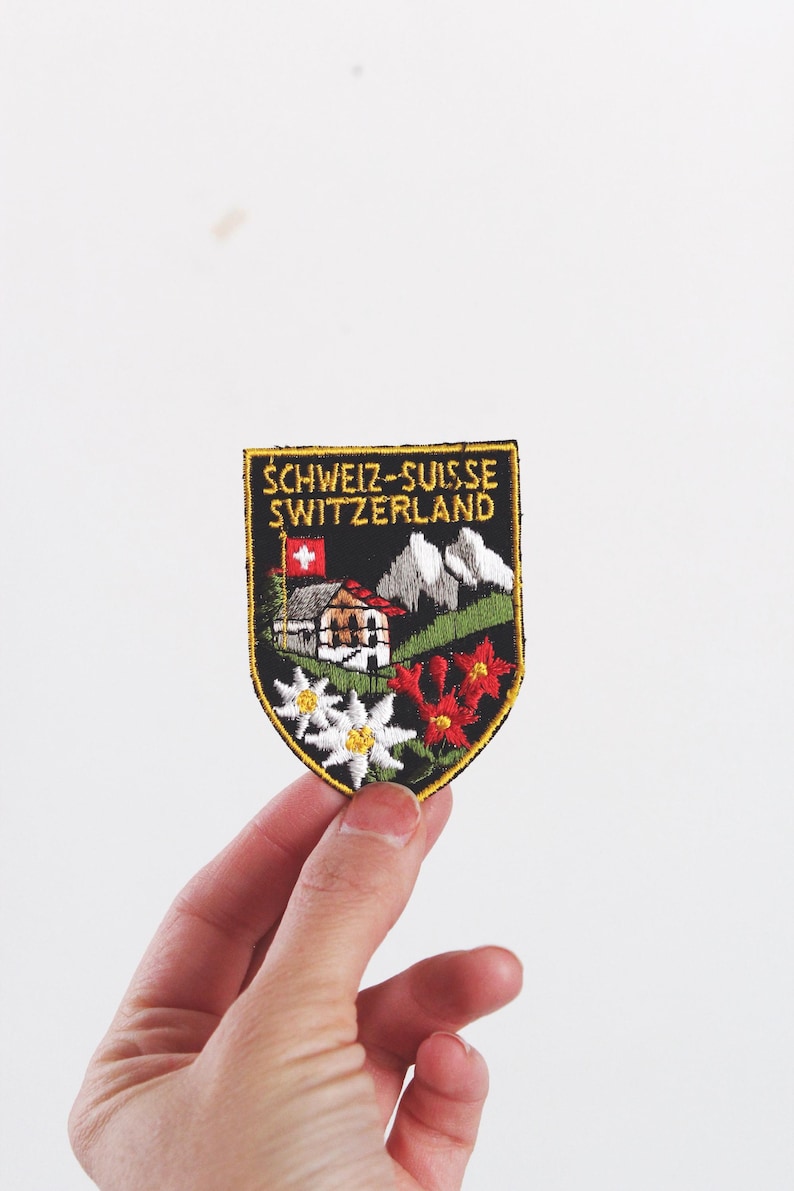 Może przedstawiać: Naszywka w kształcie tarczy z czarnym tłem i ż&oacute;łtym napisem SCHWEIZ-SUISSE SWITZERLAND. Wz&oacute;r przedstawia małą szwajcarską flagę, g&oacute;rski domek, ośnieżone szczyty, białe kwiaty szarotki oraz czerwone kwiaty.