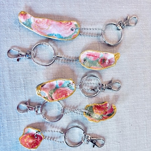 May include: Keychains with floral designs. Each keychain features a small, oblong charm with a painted floral pattern in shades of pink, red, and green. The charms are attached to a silver-colored chain and a clasp.