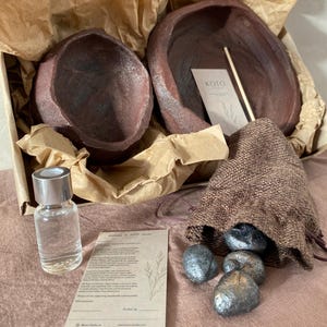 Può includere: Un set di due ciotole in ceramica marrone scuro dalla superficie ruvida, inserite in una scatola di cartone con carta marrone. Include una piccola bottiglia in vetro trasparente con tappo argentato, un sacchetto in tessuto marrone e due cartoncini.