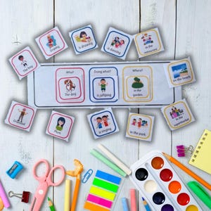 May include: Educational flashcards with illustrations and text, including phrases like "The dog is jumping" and "in the garden." The cards are arranged on a white surface with colorful art supplies, such as scissors, crayons, and watercolors.