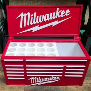 Puede incluir: Un contenedor rojo tipo caja de herramientas con el logotipo "Milwaukee" en blanco. El contenedor tiene una tapa con bisagras y un interior con múltiples compartimentos circulares y un espacio rectangular más grande. La caja de herramientas está diseñada para guardar herramientas.