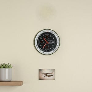 Könnte beinhalten: Eine runde Wanduhr im Aviatik-Stil mit schwarzem Zifferblatt, roten Zeigern und silbernen Details. Darunter ein gerahmtes Bild eines Doppeldeckers im Flug. Die Uhr ist an einer cremefarbenen Wand befestigt.