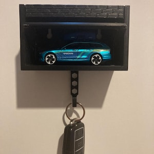 Peut inclure: Un porte-clés imprimé en 3D noir en forme de garage avec une voiture jouet bleue à l'intérieur et une clé de voiture noire suspendue à un crochet.