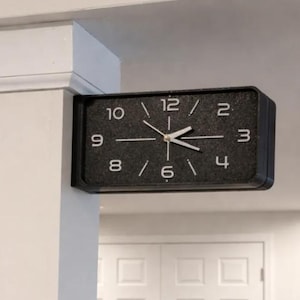 May include: A rectangular wall clock with a black textured face and white numerals 2, 3, 4, 6, 8, 9, 10, and 12. The clock features white hour, minute, and second hands mounted in the center, attached to a white pillar extending from the wall.