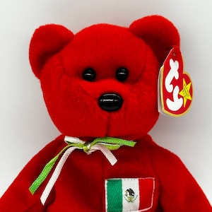 May include: Red plush teddy bear with a green and white ribbon around its neck. The bear has a black nose and eyes and a small embroidered Mexican flag on its chest. The bear has a red Ty tag with a white star.