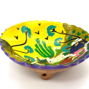 Pode incluir: Tigela de cerâmica pintada à mão com interior amarelo apresentando um desenho de paisagem com árvores, pássaros e figuras. A borda é roxa e a base é de cor terracota natural. A tigela tem uma borda ondulada.