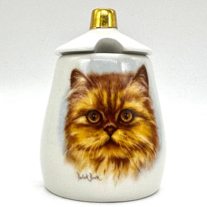 May include: White ceramic sugar dispenser with a gold lid and a painted image of a brown and orange Persian cat. The cat's eyes are green and the image is signed "Derrick Boon".