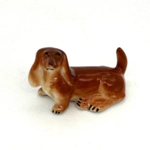 Puede incluir: Una pequeña figurita de dachshund de cerámica marrón. La figurita está tumbada con la cabeza girada hacia un lado y las orejas caídas.