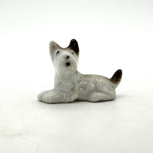 Puede incluir: Pequeña figura de cerámica de un perro, probablemente un Scottish Terrier, en posición de descanso. El perro es principalmente blanco con detalles marrones en las orejas y la cola. La figura es detallada y parece vintage.