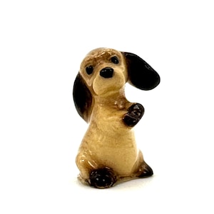 Puede incluir: Pequeña figura de perro salchicha de cerámica en posición sentada. El perro tiene un cuerpo marrón claro, orejas negras y patas negras. La figura está detallada con un acabado brillante y una expresión amigable.