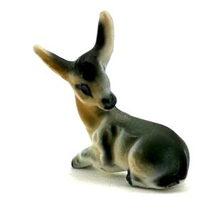 Puede incluir: Pequeña figura de ciervo de cerámica en posición de descanso. El ciervo tiene un cuerpo de color beige claro con marcas grises y negras más oscuras. Sus orejas están erguidas y tiene una expresión suave. Sobre fondo blanco.