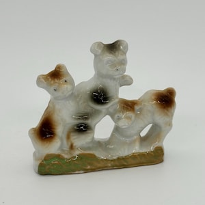 Puede incluir: Una figurita de porcelana de tres perros marrones y blancos juguetones. Los perros están de pie sobre una base verde y marrón.