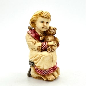 Puede incluir: Una figura de un niño con una túnica color crema, sosteniendo un pequeño oso de peluche. La túnica tiene adornos rojos y el niño tiene el pelo rubio. La figura está sentada en una silla.