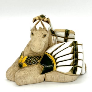 Puede incluir: Una figurita de cerámica de un caballo con un tocado con detalles en blanco, negro y amarillo. El caballo está acostado con la cabeza girada hacia un lado.