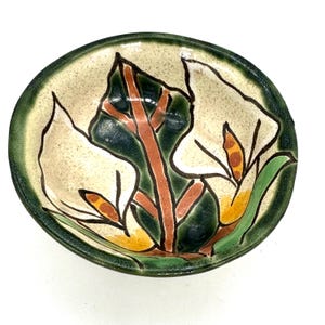 Pode incluir: Uma tigela de cerâmica pintada à mão com um design floral. A tigela apresenta um interior creme e uma borda verde. O design floral inclui copo-de-leite brancos com folhas verdes e caules castanhos. A tigela tem aproximadamente 15 cm de diâmetro.