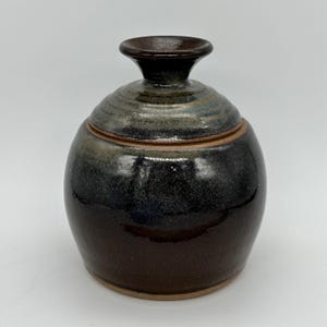 Może przedstawiać: Mały, okrągły ceramiczny słoik z pokrywką. Słoik jest pokryty glazurą w odcieniach brązu, czerni i szarości. Pokrywka ma mały guzik na górze.