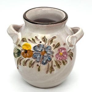Puede incluir: Un pequeño jarrón de cerámica blanquecina con dos asas y un borde marrón. El jarrón está decorado con diseños florales coloridos en relieve en amarillo, azul y rosa. Las flores están rodeadas de hojas verdes.