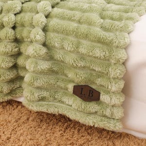 May include: A sage green plush throw blanket with a textured, ribbed design. A white teacup and saucer sit on an open book. The blanket has a brown leather tag with the initials "LB".