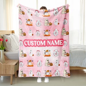May include: Pink Halloween-themed blanket featuring repeating ghost illustrations with pumpkins and soda cans. The blanket has the text "CUSTOM NAME" in a pink banner. The blanket is made of a soft material.