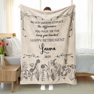 May include: A beige blanket with black floral and botanical illustrations. The blanket has the text "NEVER UNDERESTIMATE the difference YOU MADE OR THE lives you touched HAPPY RETIREMENT Laura 2025".
