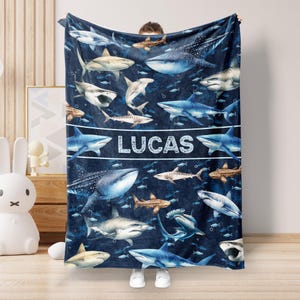 May include: A blue fleece blanket with a shark pattern and the name "LUCAS" printed in white. The blanket features various types of sharks, including hammerhead, tiger, and whale sharks.