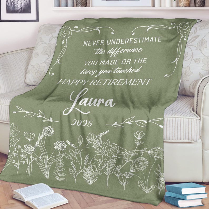 May include: A sage green throw blanket with white floral designs and text. The text reads "NEVER UNDERESTIMATE the difference YOU MADE OR THE lives you touched HAPPY RETIREMENT Laura 2025". The blanket is draped over a sofa.