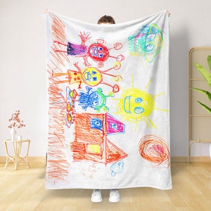 Könnte beinhalten: Eine weiße Fleece-Decke mit einer bunten Zeichnung eines Hauses, einer Sonne und Außerirdischen. Die Zeichnung ist mit Buntstiften gemacht und sieht aus wie ein Kinderbild.