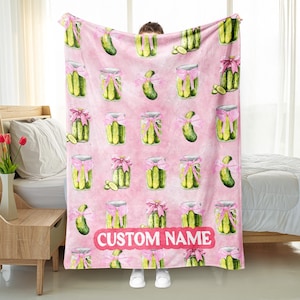 May include: A pink blanket featuring a pattern of jars of pickles tied with pink bows and whole pickles. The blanket has the text "CUSTOM NAME" in a pink banner at the bottom.