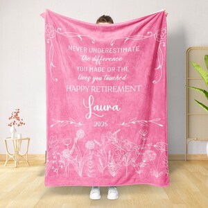 May include: A vibrant pink blanket with white floral accents and text. The text reads "NEVER UNDERESTIMATE the difference YOU MADE OR THE lives you touched HAPPY RETIREMENT Laura 2025". The blanket is a soft, cozy textile.