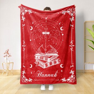 May include: Red fleece blanket with a white design of a dragon flying over a stack of books. The design also includes stars, moons, and the name "Hannah".