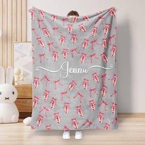 Könnte beinhalten: Graue Decke mit einem Muster aus roten Schleifen und Coca-Cola-Dosen. Der Name "Jenna" ist in weißer Schrift quer über die Mitte geschrieben. Eine weiche und gemütliche Decke.