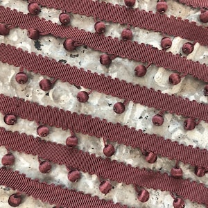 May include: Close-up of maroon fringe trim with small, round, decorative bobbles. The trim has a ribbed texture and is arranged diagonally across a textured, light-colored surface. This decorative trim is suitable for various craft and sewing projects.