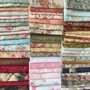 May include: A collection of folded fabric pieces in various colors and patterns. The fabrics include floral, striped, and textured designs in shades of pink, blue, green, and cream. The image showcases a variety of textiles suitable for sewing and crafting projects.