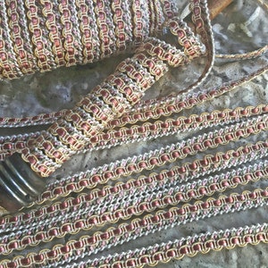 May include: A spool of gold and rose gold trim with a white and pink woven pattern. The trim is arranged in a decorative pattern on a gray surface.