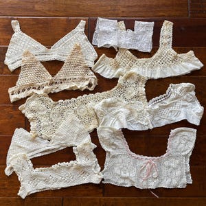 May include: A collection of antique, off-white crochet tops and bodice pieces. The handmade garments feature intricate lace patterns, varying necklines, and some with ruffled edges. The pieces are arranged on a dark wood surface, showcasing the delicate craftsmanship.