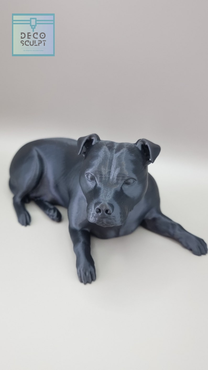 May include: A black 3D printed figurine of a dog lying down. The dog has a short, smooth coat and a short tail. The figurine is detailed and realistic.