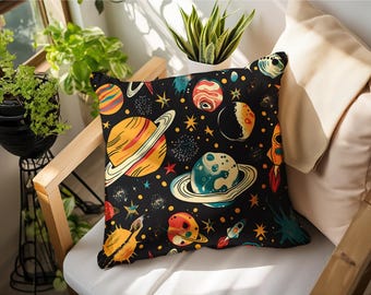 May include: A square throw cushion featuring a black background with a colourful space-themed pattern. The design includes various planets with rings, stars, and small rockets in shades of orange, yellow, blue, and red across the fabric surface.