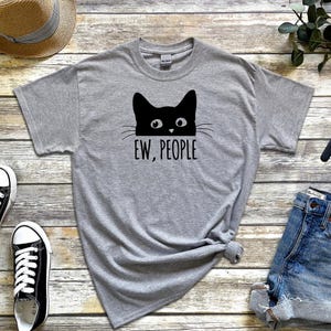 Pu&ograve; includere: Maglietta grigia con un gatto nero che spunta da sopra e la scritta "EW, PEOPLE" stampata sotto.